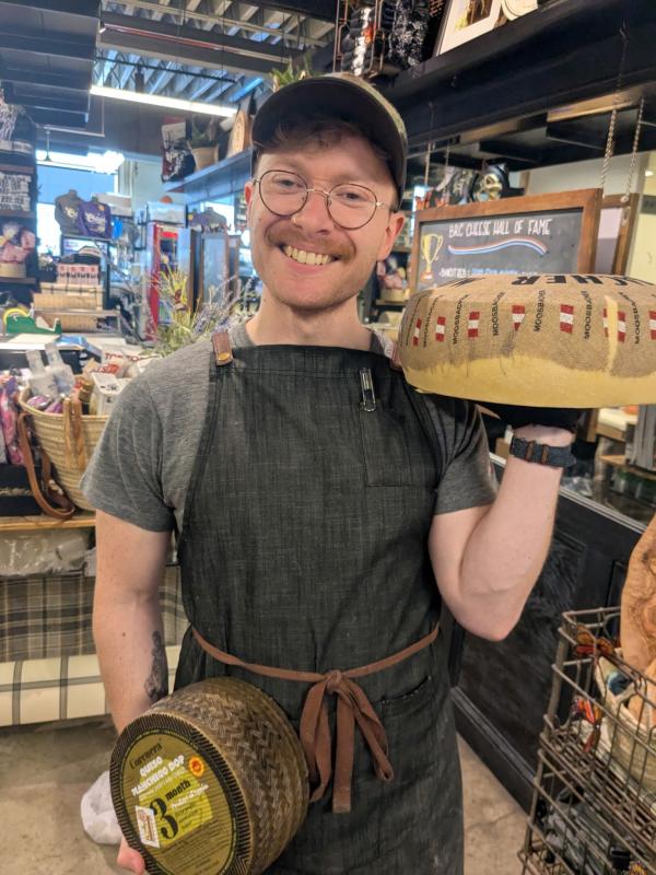 Man with glasses, apron, mustache holding two large wheels of cheese